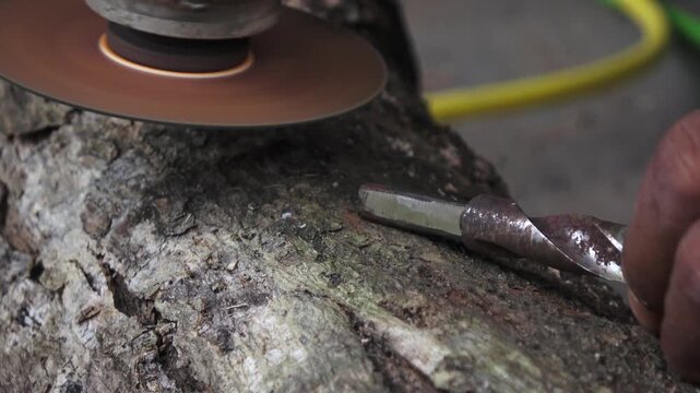 A high-quality close-up shot focusing on the precise process of sharpening a metal drill bit using a grinding tool. The scene is set outdoors against a rustic teak wood log, highlighting industrial ma