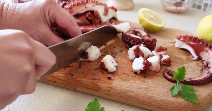 Woman hands slicing cooked octopus with sharp chef knife on wooden board. Person preparing fresh seafood salad with lemon and parsley for healthy mediterranean meal. Culinary process of cutting boiled