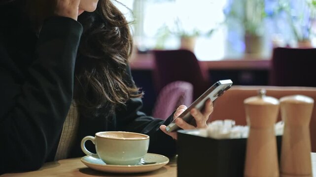 Young woman using mobile phone and texting in social networks, sitting in coffee shop. Lady drinking cappuccino while swiping on phone screen. Lifestyle concept. Real time