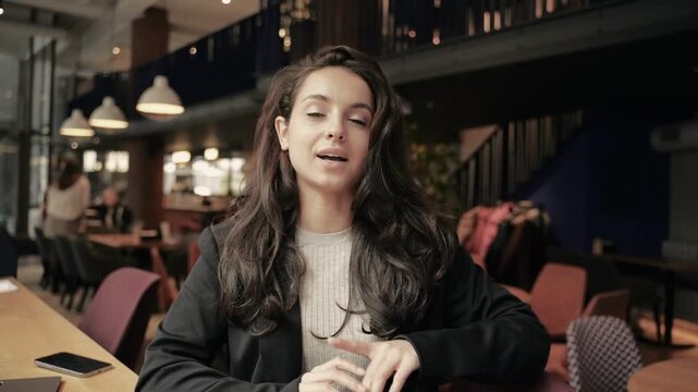 Happy young woman blogger sitting in restaurant and looking at camera. Lady doing online job interview during video chat conference call. E-coaching, online training concept. Real view