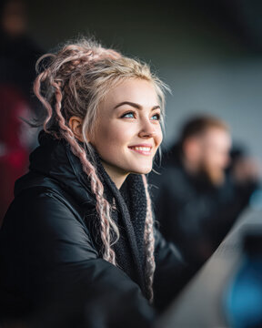 A young woman with pink dreadlocks smiles while sitting indoors, bundled in a black jacket, with a blurred background of people.