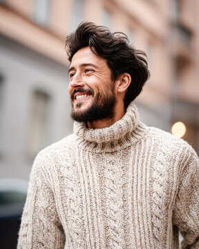 A smiling man with a beard wears a cozy knitted sweater while standing outdoors in an urban setting.