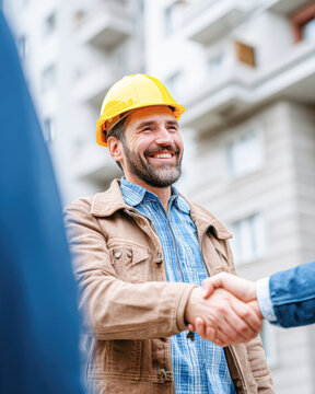 A smiling construction worker in a yellow hard hat shakes hands with another person in front of a building.