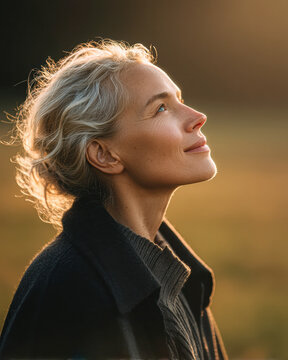 A serene woman with silver hair gazes thoughtfully upward, bathed in warm, golden sunlight outdoors.