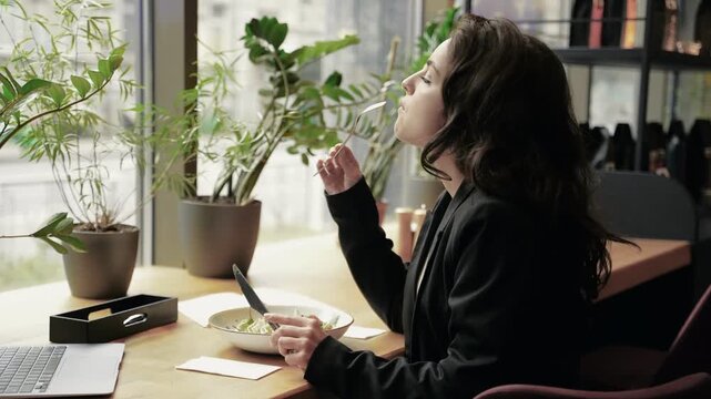 Happy young woman eating salad at restaurant while looking out the window at the street. Smiling lady having lunch in cafe during break. Healthy food concept. Real time