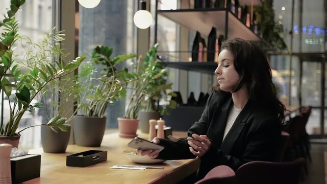 Happy attractive lady eating salad and talking on smartphone at cafe. Smiling young female taking a phone call during lunch in restaurant. Healthy food concept. Real time