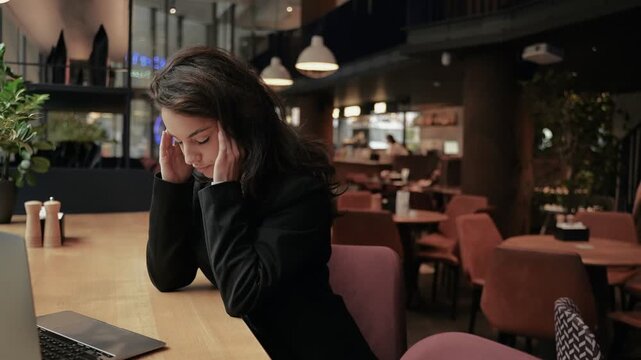 Tired young businesswoman working online and using laptop in cafe. Keeping hands on head, massaging of temples. Headache, migraine. Real time