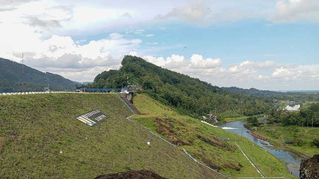 A scenic view of a tropical green hillside with a small dam and flowing river, surrounded by lush vegetation under a bright blue sky with clouds. Ideal for nature, travel, environment, and outdoor the