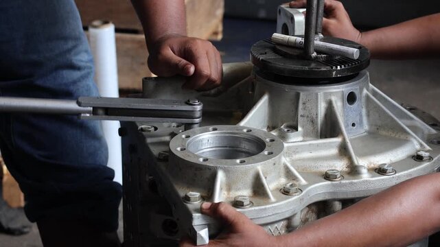 A medium static shot showing mechanics working together to assemble or repair a heavy industrial gearbox. One technician is using a torque wrench.