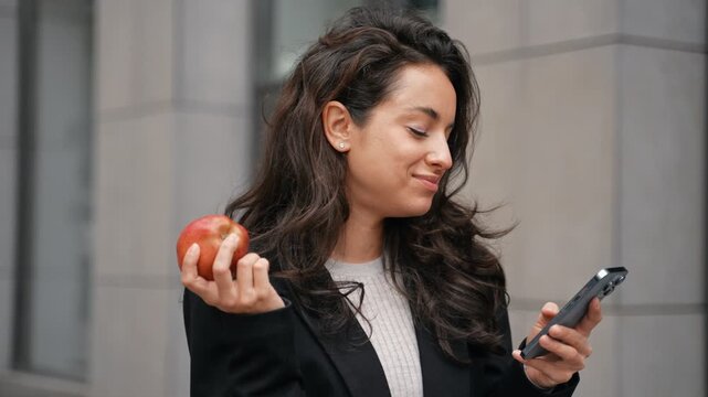Businesswoman biting an apple while holding smartphone and looking at the screen. Young lady standing outdoor and using mobile phone. Lifestyle, food concept. Real time