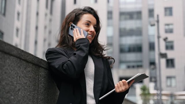Happy pretty woman talking on smartphone outdoors. Lady standing near wall and holding documents, digital tablet. Businesswoman working online. Real time