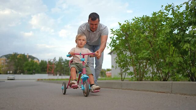 Happy father pushes little son on tricycle outdoors. Dad helps small boy ride bike on path. Family walks near green bushes. Father cares for child during summer day. Dad and son enjoy time.