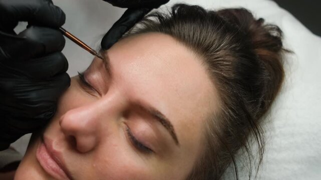 Close-up of a brow artist applying henna with a brush during a permanent makeup procedure in a salon