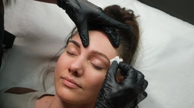 Professional beautician applying permanent makeup and cleaning a woman's eyebrows in a beauty salon