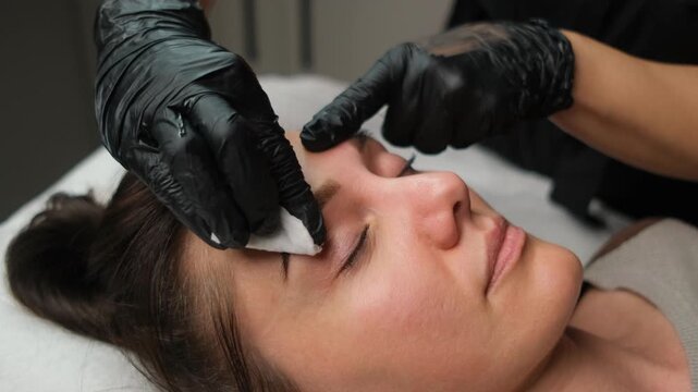 Cosmetologist cleaning a woman's eyebrows with a cotton pad after a microblading treatment