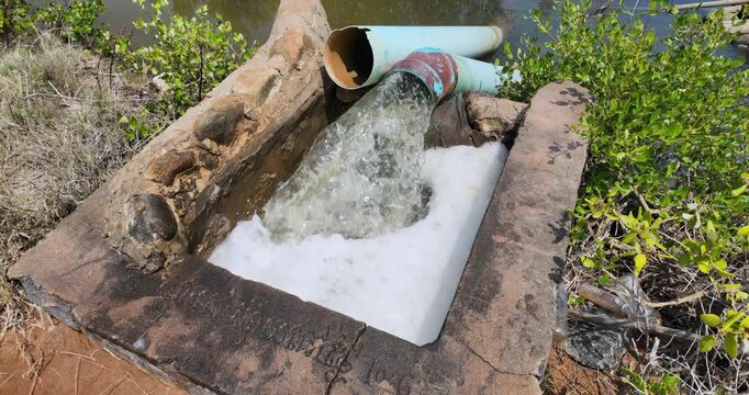 close up of a rural pumping station using a diesel engine to discharge water from blue pipes, creating white foam and bubbles in a stone lined basin.