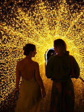 Two women walking through glowing golden light tunnel at night, immersive illuminated pathway and magical experience concept