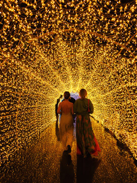 Two women walking through glowing golden light tunnel at night, immersive illuminated pathway and magical experience concept