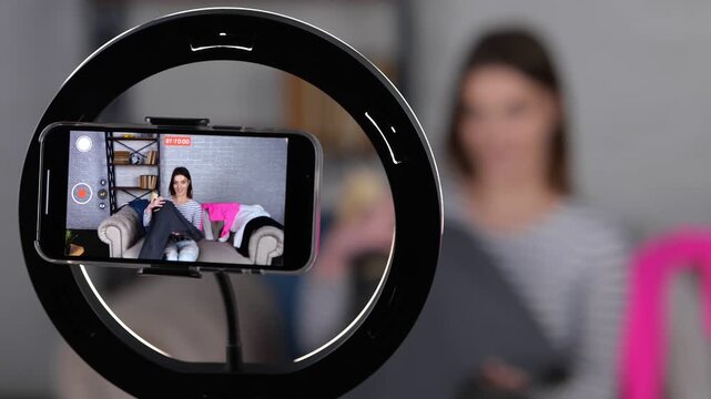Smiling female blogger sitting on sofe, using smartphone while recording video for her vlog, selling online and showing jumper. Small business owner, online market concept. Real time video.