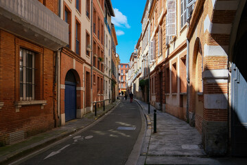 Fototapeta premium Narrow cobblestone street in Toulouse, France, featuring traditional red brick architecture and shutters under a clear blue sky. Historic European city alley with old buildings and pedestrian path