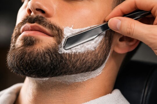 A man"s beard is being shaved with a straight razor, removing shaving cream for a clean, groomed look.