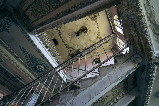Decayed abandoned staircase viewed upward, ornate railings, peeling plaster, historic building interior, urbex, eerie architectural geometry.