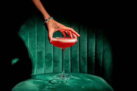 Hand reaching for a red cocktail with foam in a coupe glass on a green velvet chair. High-contrast dramatic lighting, luxury bar concept
