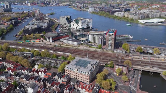 Amsterdam cityscape flying over traditional dutch houses, showing IJ river waterway, modern architecture, railroad tracks leading to Centraal Station in Netherlands