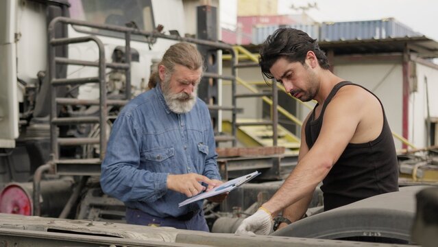 A senior mechanic supervises a younger technician performing truck maintenance, collaborating on a vehicle inspection for fleet management at a logistics depot.