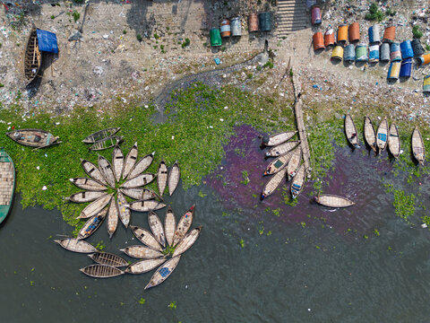 Aerial view of wooden boats arranged in flower patterns on the Buriganga River with water hyacinths and colorful barrels on the shore in Dhaka, Dhaka Division, Bangladesh.