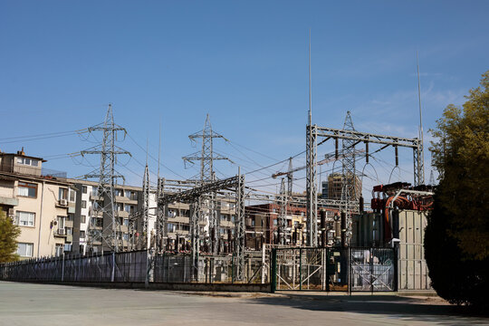 High voltage electric substation with transmission pylons and transformers located near residential apartment buildings in urban area, blue sky