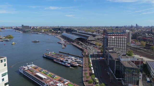 Flying sideways revealing modern buildings, Amsterdam Centraal station, bustling city center, tour boats sailing on IJ river during sunny day
