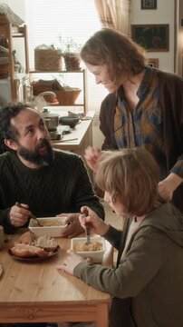 Vertical shot of woman serving food, placing meals on table, family sitting and eating together in small apartment
