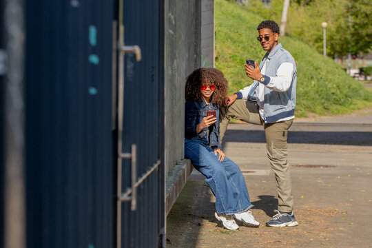 Two young stylish friends relaxing outside, smiling and using smartphones. Urban setting, sunny day, casual denim outfits, positive vibes and modern lifestyle