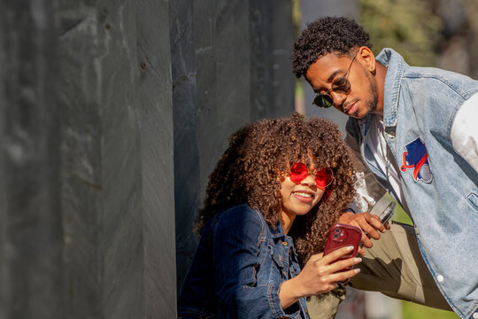 Two young stylish friends relaxing outside, smiling and using smartphones. Urban setting, sunny day, casual denim outfits, positive vibes and modern lifestyle