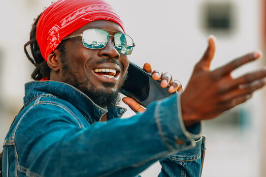Smiling young man with dreadlocks, red bandana and mirror sunglasses talking on smartphone, gesturing expressively, wearing denim jacket outdoors