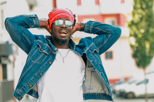 Cheerful young African man laughing outdoors, wearing denim jacket, red bandana and mirrored sunglasses, hands behind head, urban background