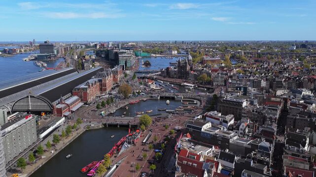 Amsterdam Centraal Station aerial shot revealing Damrak waterfront with city canals, busy streets, historic architecture under sunny sky with boats moving on water