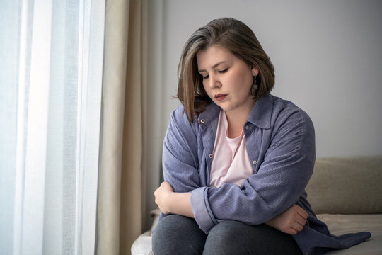 Lonely plus size woman sitting on bed by window, emotional struggle, anxiety, depression concept, mental health awareness, body positivity.