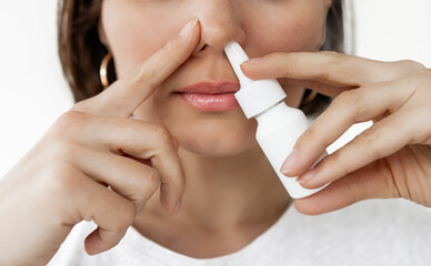 Fototapeta premium Brunette Woman Using Nasal Spray While Closing One Nostril with Finger on White Background, Close-up Portrait of Female Treating Nasal Congestion, Allergy or Cold Symptoms
