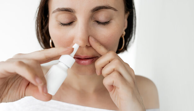 Brunette Woman Using Nasal Spray While Closing One Nostril with Finger on White Background, Close-up Portrait of Female Treating Nasal Congestion, Allergy or Cold Symptoms