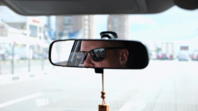 Eyes in sunglasses of male driver reflecting in rearview mirror while riding motor auto. Young guy looking concentrated to urban road during operating vehicle. Confident man driving modern car at city