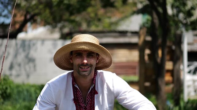 Portrait of ukrainian man in straw hat against background of summer garden. Young smiling guy wearing an embroidered shirt looking into camera outdoor. Concept of national identity and patriotism