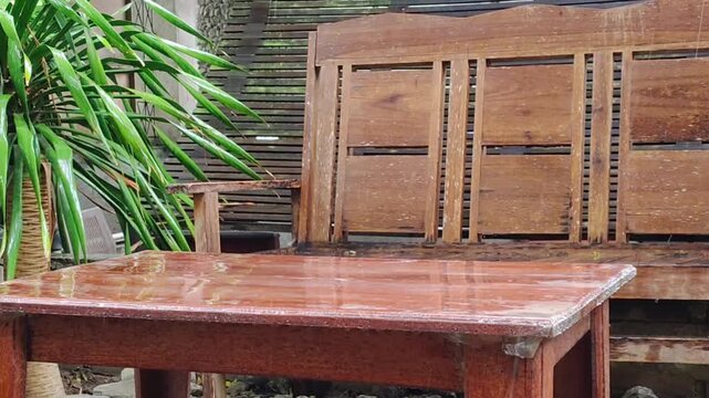 Raindrops falling and splashing on a wet wooden table and empty bench beside a green plant in a peaceful outdoor garden.