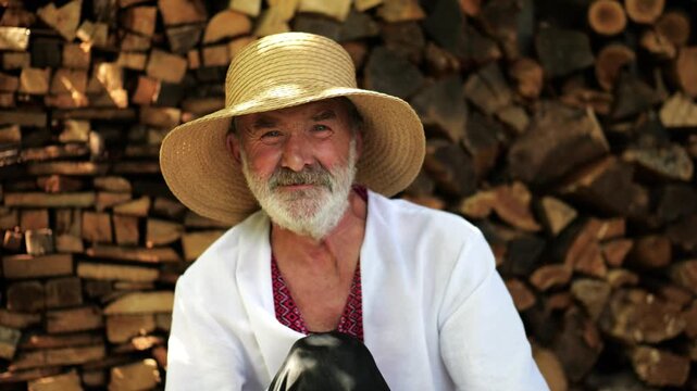 Older smiling male wearing an embroidered shirt looking into camera outdoor. Portrait of senior ukrainian man in straw hat against background of woodshed. Cultural heritage and ethnic roots concept