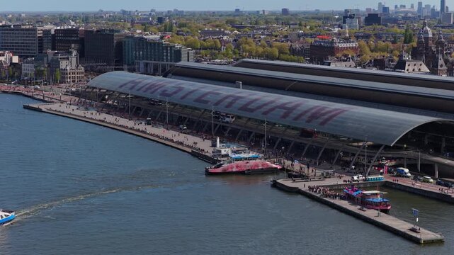 Aerial shot of Amsterdam Centraal station showcasing ferries navigating IJ river waters with cityscape, historic architecture, urban transportation network in Netherlands