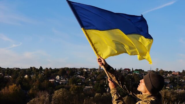 Ukrainian army man stands with raised national banner to peak of hill. Male soldier in military uniform with Ukraine flag as symbol of victory against russian aggression. Invasion resistance concept