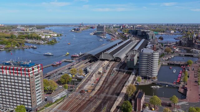 Aerial perspective of Amsterdam Centraal station showcasing extensive railway infrastructure and bustling river traffic on IJ with cityscape panorama during sunny day