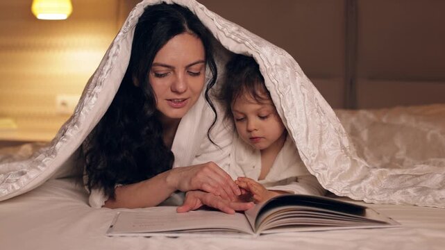 Loving mother reading a book to her little daughter in bed at night. Happy family enjoying a bedtime story, creating warm memories together