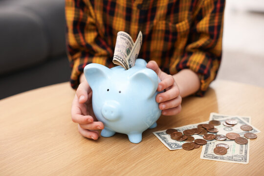 Little boy with piggy bank and money at wooden table indoors, closeup